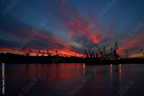 Colorful sunset over sea port and industrial cranes. Bright sunset in seaport. Large silhouettes of cargo cranes. Beautiful landscape with fiery sunset sky and sea.