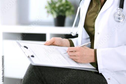 Woman doctor writing something at clipboard while sitting at the chair  close-up. Therapist at work filling up medication history records. Medicine and healthcare concept