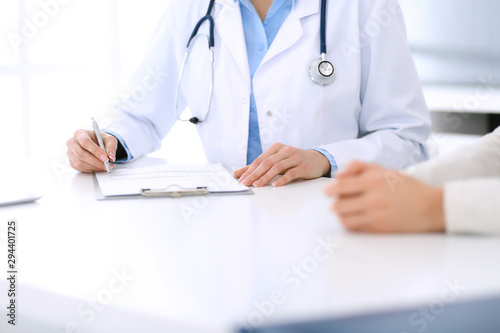Woman doctor and patient sitting and talking at medical examination at hospital office, close-up. Physician filling up medication history records. Medicine and healthcare concept