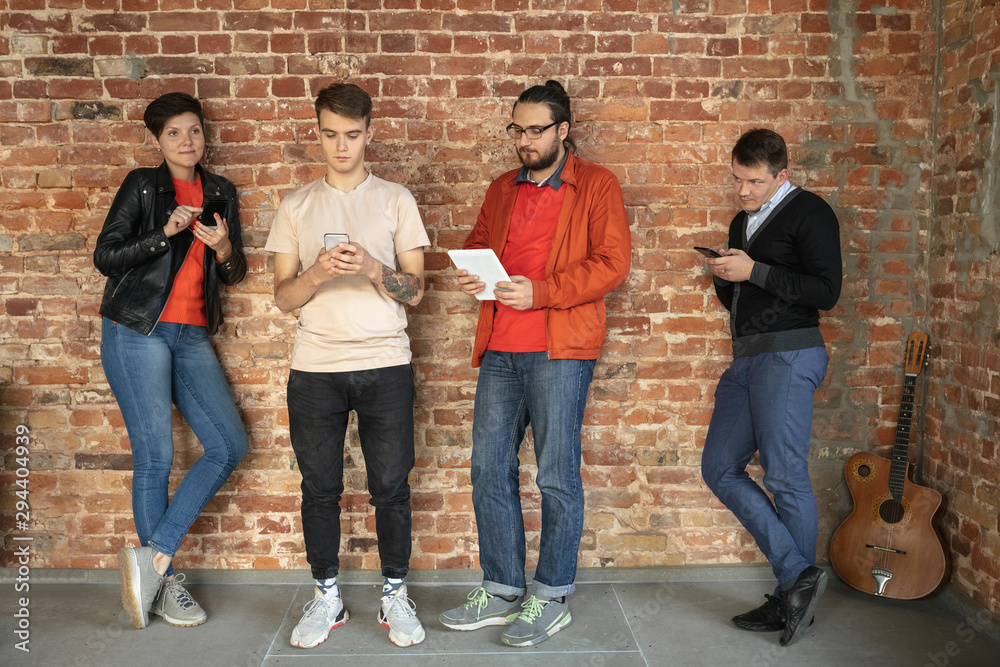 Fototapeta premium Group of happy caucasian young people standing behind the brick wall. Sharing a news, photos or videos from smartphones or tablets, playing games and having fun. Social media, modern technologies.