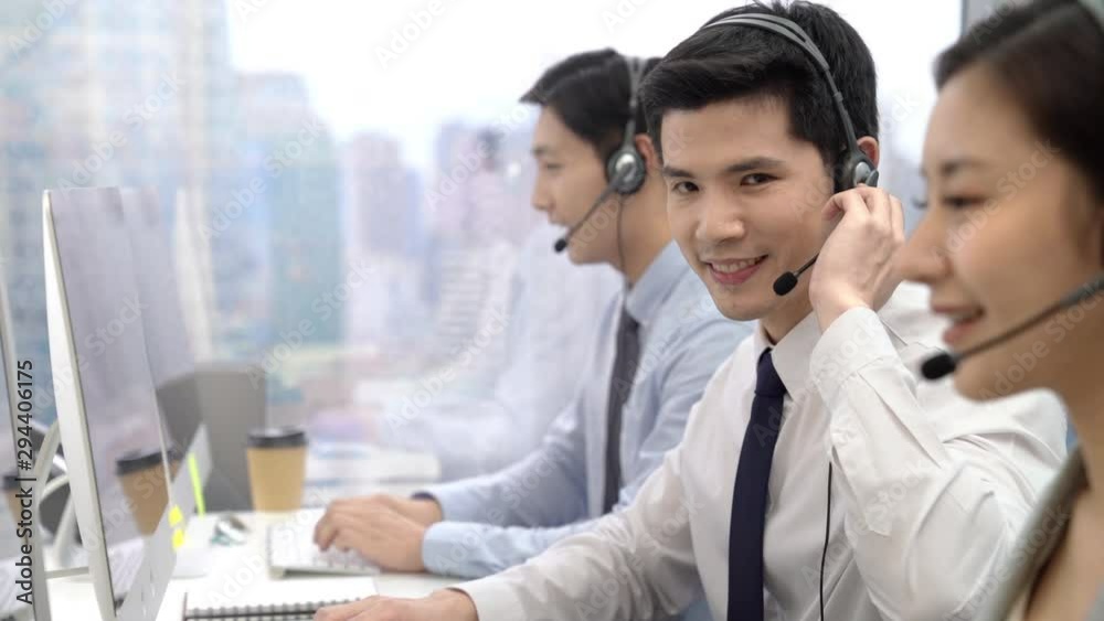 Young Asian male telemaketing operator smiling to the camara while working at call center office in the city