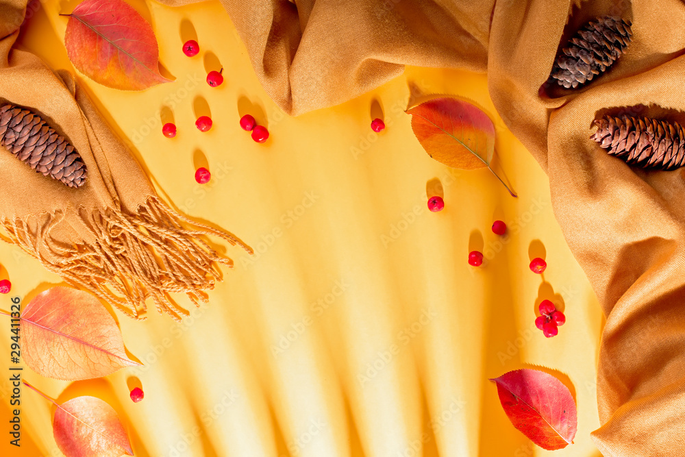 Fototapeta premium Autumn composition with cozy soft yellow scarf or blanket, leaves, red berries, pine cones on sunny yellow background. Fall border with autumn fashion accessories and orange leaves, top view