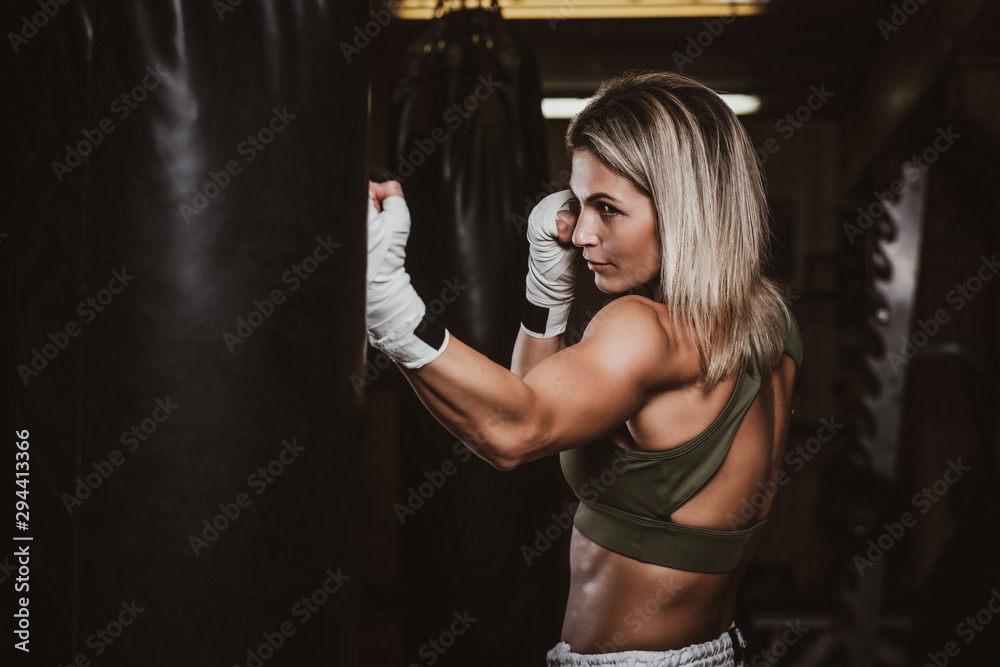 Muscular pretty woman is doing her kickboxing exercises with punching bag.