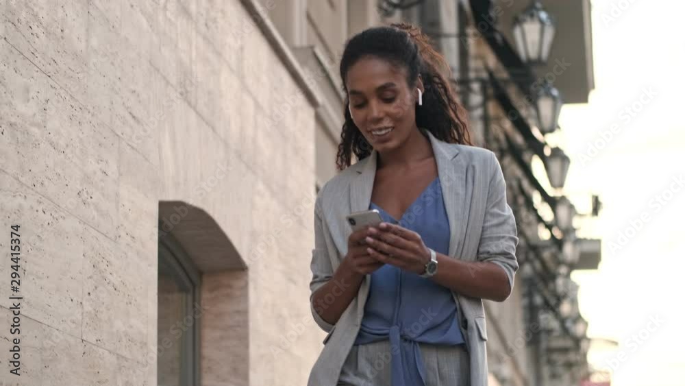 Bottom view of happy young african business woman listening music with earpods on smartphone while walking at the street Bottom view of happy young african business woman listening music with earpods on smartphone while walking at the street