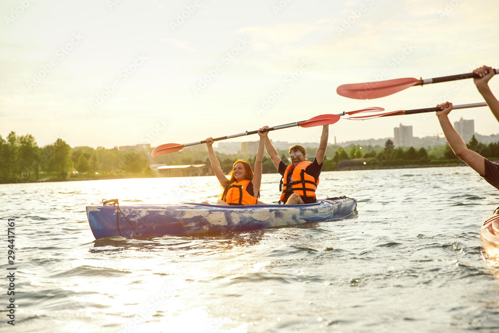 Obraz premium Happy young caucasian couple kayaking on river with sunset in the backgrounds. Having fun in leisure activity. Happy male and female model laughting on the kayak. Sport, relations concept. Colorful.