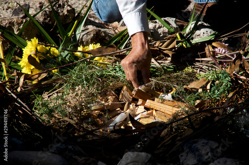 A mayan priest is placing lit candles and wood on ceremonial fire ritual, flowers and herbs can be seen.