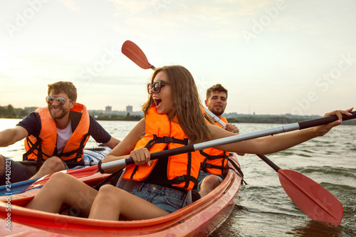Happy young caucasian group of friends kayaking on river with sunset in the backgrounds. Having fun in leisure activity. Happy male and female model laughting on the kayak. Sport, relations concept.