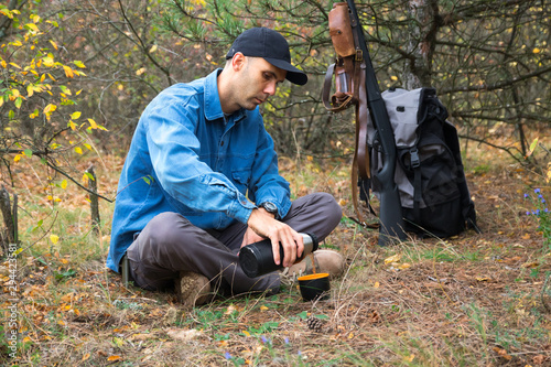 caucasian male hunter resting in forest. drinking tea from thermos. shotgun, ammunition, backpack on the background