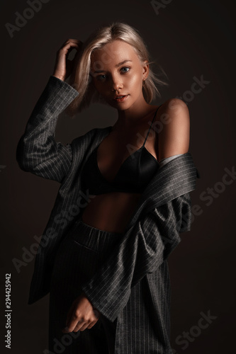 Stylish fashion trendy blonde girl with elegant hairstyle in classic trousers and black lingerie posing on black background. Beautiful hot sexy portrait of young woman model underwear in room
