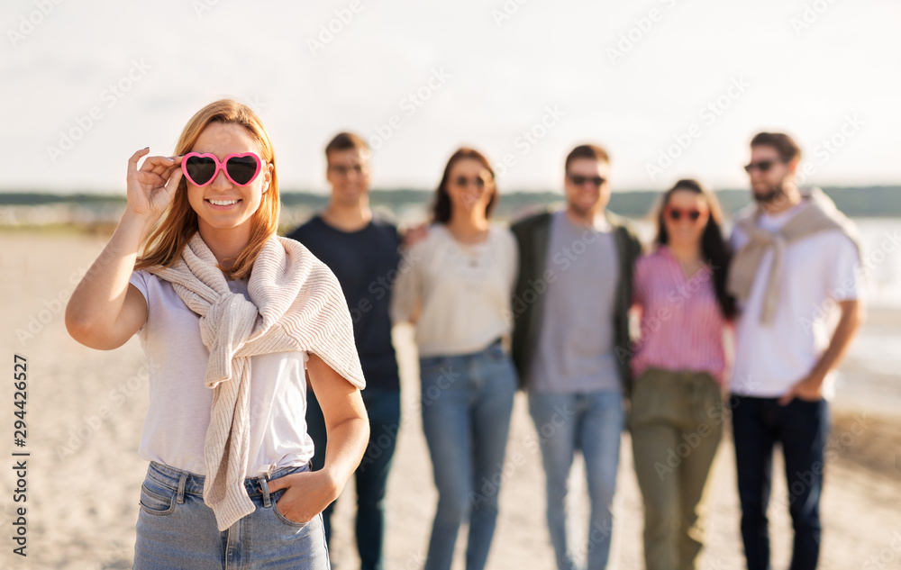 Obraz premium friendship, valentine's day and people concept - happy woman in heart-shaped sunglasses with group of friends on beach in summer