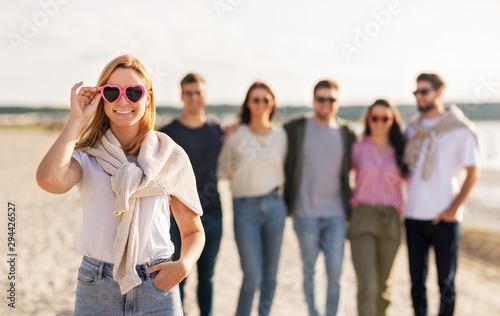 friendship, valentine's day and people concept - happy woman in heart-shaped sunglasses with group of friends on beach in summer