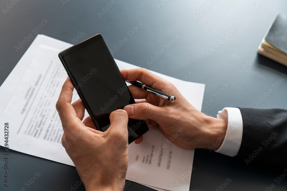 Top view of businessman hands in dark suit holding smartphone with blurred office table with contract in background. Concept of internet and technology Top view of businessman hands in dark suit holding smartphone with blurred office table with contract in background. Concept of internet and technology