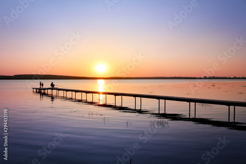 Colorful sunset over the lake with a pier. A fisherman is fishing on the pier and the children came to see. Pink and purple pastel watercolor soft tones.