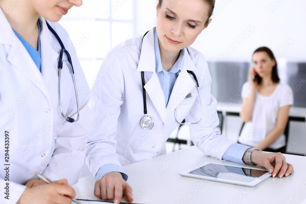 Group of doctors at work in hospital with patient in a queue at the background. Physician filling up medical documents or prescription while standing at reception desk. Data in medicine and health Group of doctors at work in hospital with patient in a queue at the background. Physician filling up medical documents or prescription while standing at reception desk. Data in medicine and health