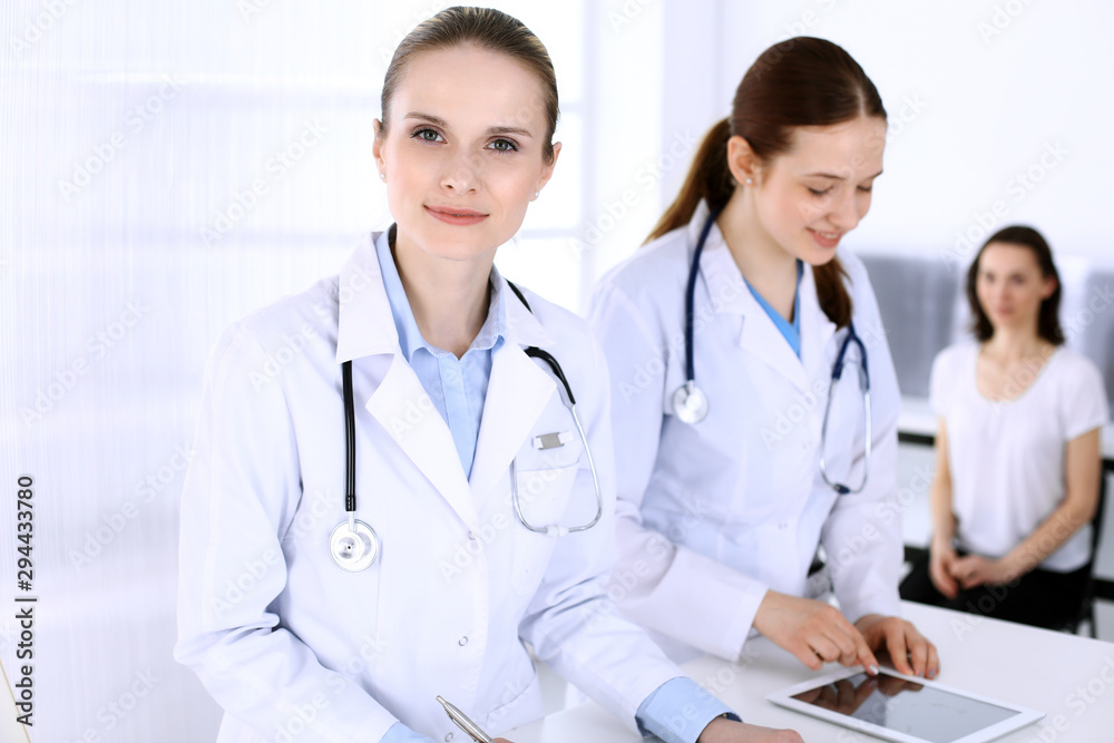 Group of doctors at work in hospital with patient in a queue at the background. Physician filling up medical documents or prescription while standing at reception desk. Data in medicine and health Group of doctors at work in hospital with patient in a queue at the background. Physician filling up medical documents or prescription while standing at reception desk. Data in medicine and health