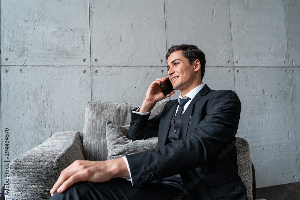 Cheerful young businessman in dark suit talking on smartphone sitting in gray armchair in loft office. Concept of communication and business lifestyle Cheerful young businessman in dark suit talking on smartphone sitting in gray armchair in loft office. Concept of communication and business lifestyle