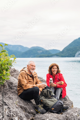 A senior pensioner couple hiking by lake in nature, sitting and resting.