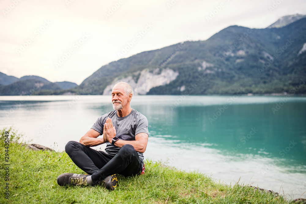 Fototapeta premium A senior man pensioner sitting by lake in nature, doing yoga exercise.