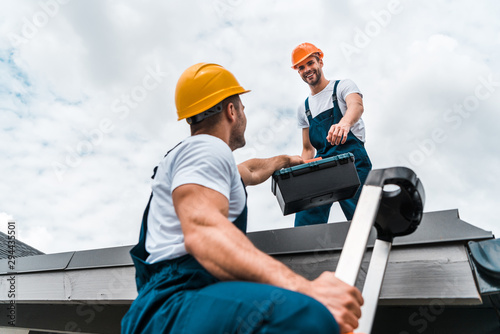 low angle view of handyman in helmet giving toolbox to happy colleague photo