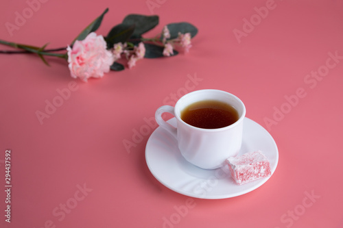 A white cup of tea with a piece of lokum, turkish delight on the side,  with delicate flowers on a pink background. Afternoon tea concept