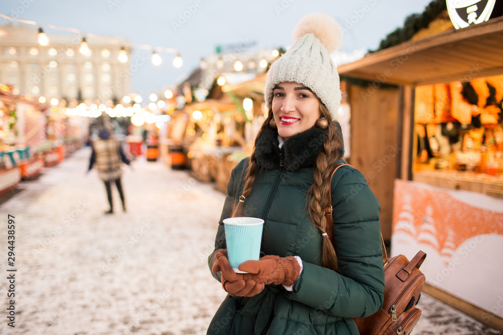Beautiful smiling girl holding a cup of tea or coffee in winter. Beautiful woman in the winter on the street with New Year decorations.