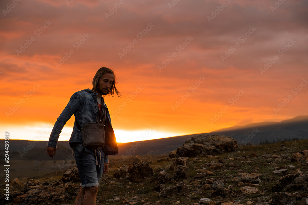 A traveler enjoys a walk in the mountains at sunset. A man in a denim jacket, shorts, long blond hair. Backpacker in a hike in the summer mountains. The sun sets over the mountains. Silhouette.
