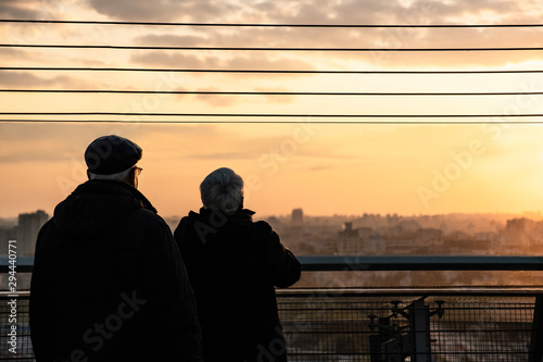 Elderly couple watching the sunset. From the roof of a tall skyscraper. The city in the rays of the setting sun. Grandpa and Grandma. A date on the roof. Meeting and communication. Gray people.