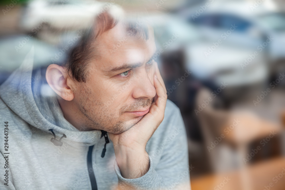 Fototapeta premium Sad pensive handsome man leaning head on hand in cafe, looking through a window with reflections.