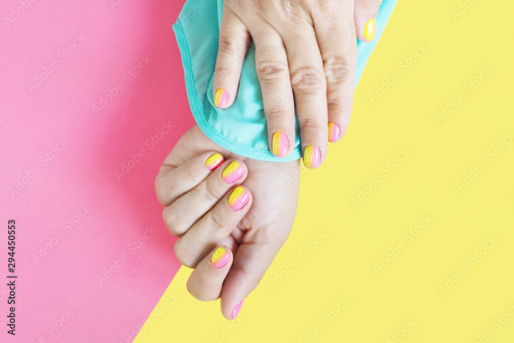 Fototapeta premium Female hands in a blue blouse with a bright pink-yellow manicure on a yellow-pink background...