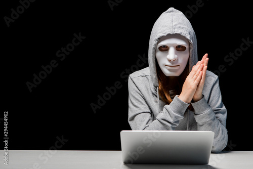 front view of anonymous girl in mask and hoodie sitting near laptop and rubbing hands during cyberbullying isolated on black photo