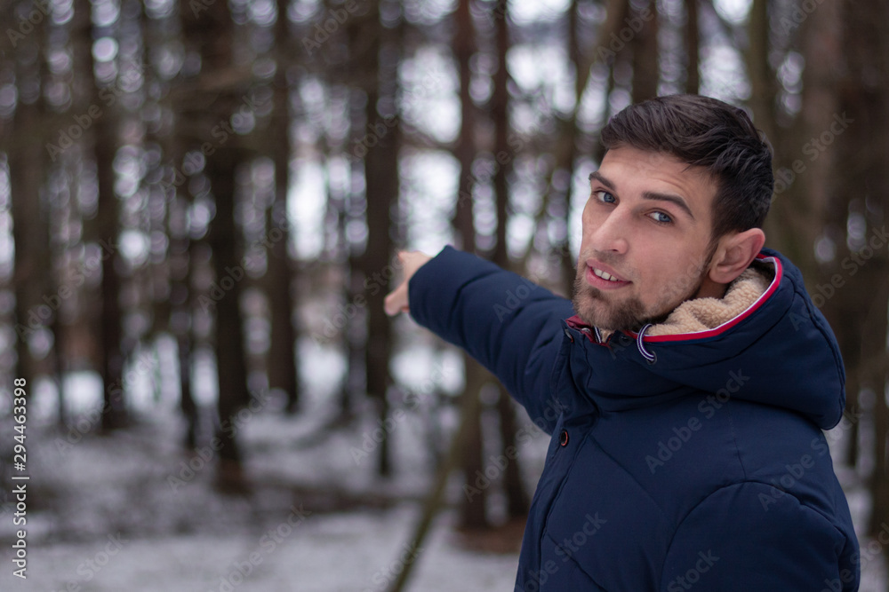 a young guy shows his index finger deep into the forest. Fashionable hairstyle, brutal bristles and a stylish beard. Dressed in a blue jacket. It's time for winter. In the background a forest, trees a young guy shows his index finger deep into the forest. Fashionable hairstyle, brutal bristles and a stylish beard. Dressed in a blue jacket. It's time for winter. In the background a forest, trees