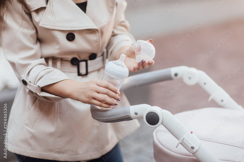 Baby food and water. Young mother is going to feed the baby with a bottle of baby drink. Happy woman walking with a stroller. Close-up young mother, pram, baby food and drink in the bottle. Baby food and water. Young mother is going to feed the baby with a bottle of baby drink. Happy woman walking with a stroller. Close-up young mother, pram, baby food and drink in the bottle.