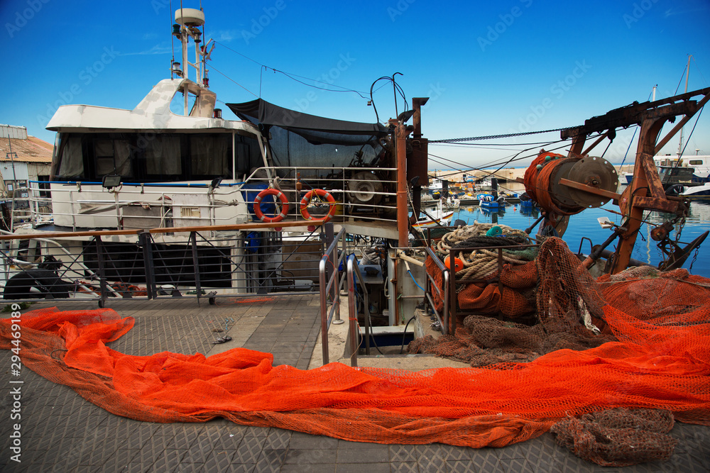 Fishing tackle on the pier. Moored fishing boat, red bright fishnet, ropes,  lifebuoys. Pier with a red fishing net in the foreground. Colorful marine  picture against a bright blue sky. Stock Photo