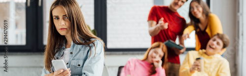 panoramic shot of group of schoolchildren laughing at sad girl with smartphone
