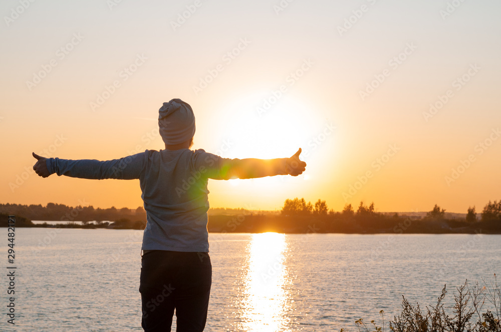 Fototapeta premium A young man, a hipster, stands on a lake at sunset, enjoys the view while traveling, desolation, enjoyment, harmony.