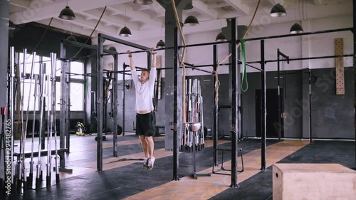 Handheld, man pulls up on bar in gym, wears lift and grip gear and white t-shirt. Fitness instructor is training, pull-ups to chest. Strengthening muscles of back, body and chest