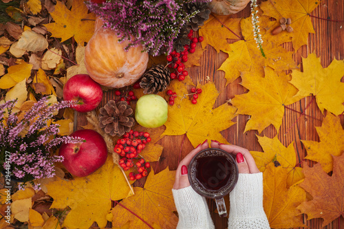 A mug of tea in a woman's hand in a sweater on the wood background with yellow falling leaves maple and cones. Hello Autumn phrase on vintage background. Hot drink. Autumn composition still life
