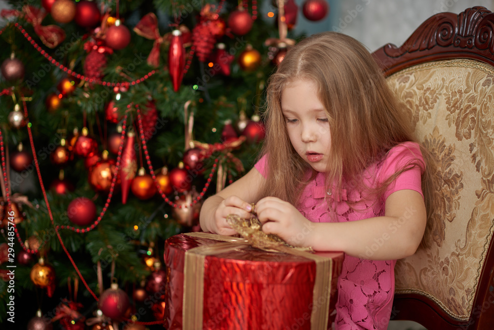 A little girl wearing light pink dress unwrapping a gift box. Decorated Christmas tree background.