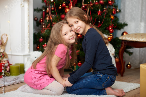 Two young sisters sitting on white carpet. Fireplace and decorated Christmas tree background. © shabbydatwin