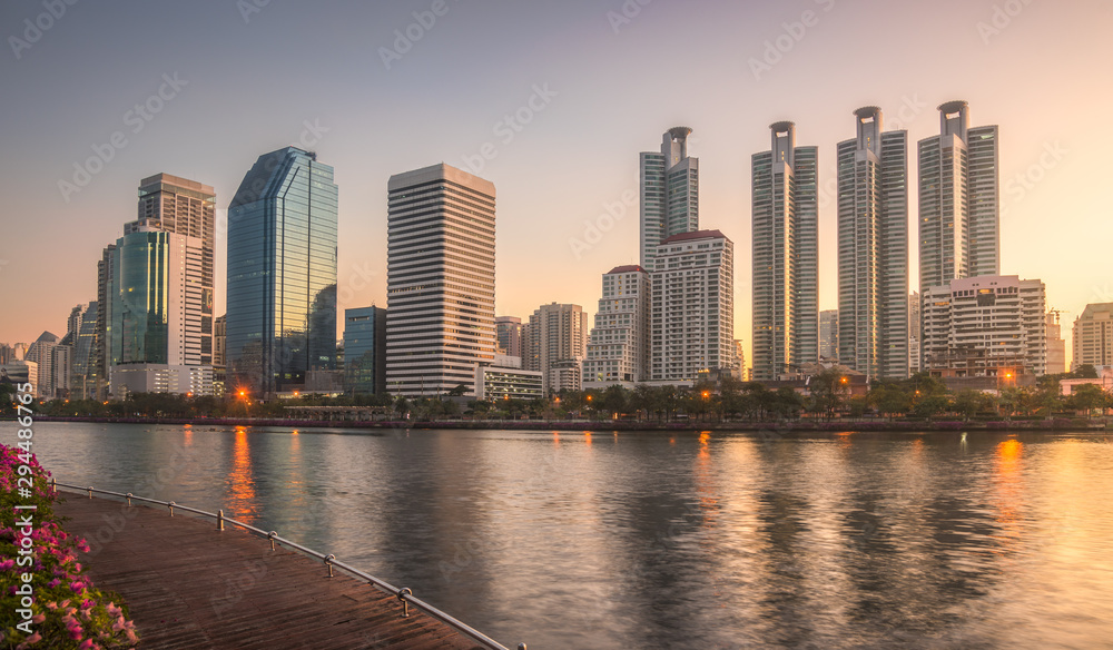 Obraz premium Skyscrapers and Lake with Wooden Walk Way in City Park. View of Benjakiti Park at Sunrise. Beautiful Morning Scene of Public Park in Bangkok, Thailand, Asia.