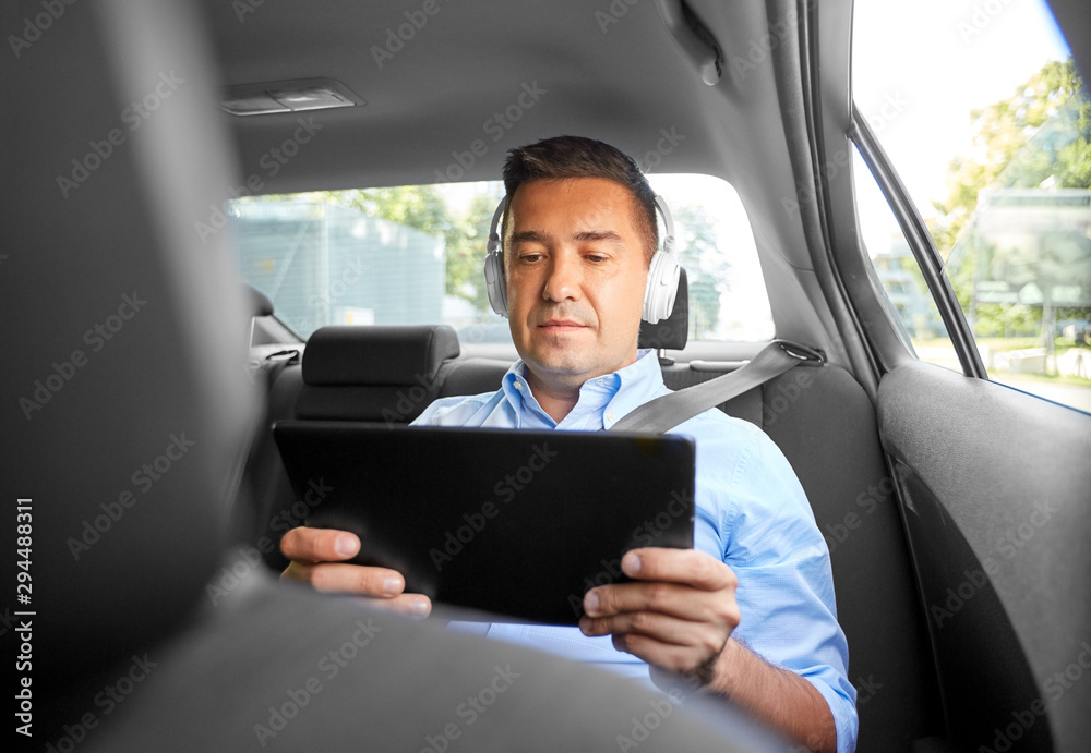 transport, business and technology concept - male passenger or businessman with wireless headphones using tablet pc computer on back seat of taxi car