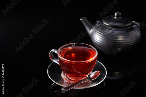 Glass cup with hot red tea and teapot on a black background.