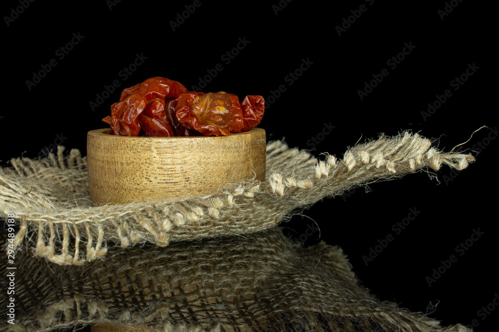 Lot of slices of dry red cherry tomato in wooden bowl on jute cloth isolated on black glass