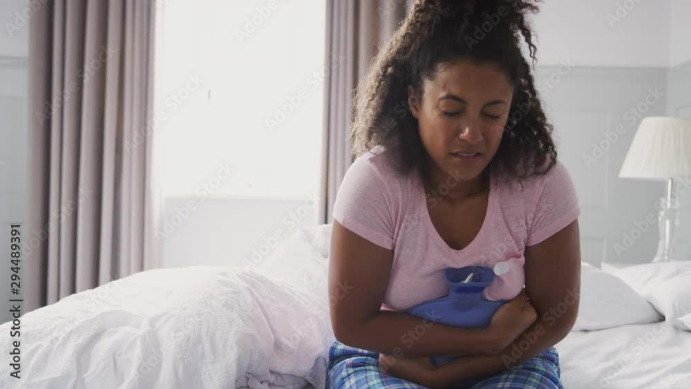 Woman Wearing Pajamas Suffering With Period Pain Sitting On Bed With Hot Water Bottle Woman Wearing Pajamas Suffering With Period Pain Sitting On Bed With Hot Water Bottle