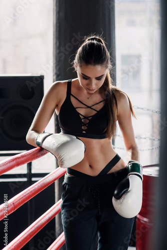 Tired sports girl leaned on red ropes on boxing ring, and have a rest after hard training in black loft gym. Healthy and sporty lifestyle concept.