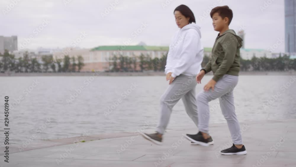 Slow motion shot of Asian mother and her young son in sportswear doing forward lunges while training together on urban riverside sidewalk on autumn day Slow motion shot of Asian mother and her young son in sportswear doing forward lunges while training together on urban riverside sidewalk on autumn day