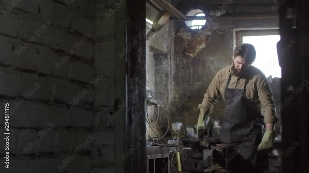 Tracking right of handsome blacksmith in safety uniform standing at anvil, holding big hammer, striking candent metal ingot with it Tracking right of handsome blacksmith in safety uniform standing at anvil, holding big hammer, striking candent metal ingot with it