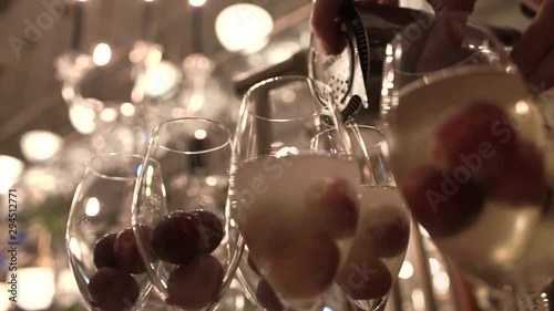 Bartender pouring cold champagne from iron shaker to tall glasses with large grapes at fancy restaurant, close-up. Skillful barman makes alcoholic cocktails on bar counter under soft evening lighting.