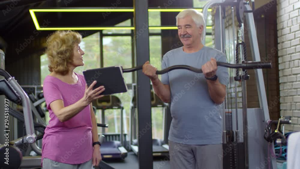 Medium shot of senior man in sportswear standing and doing exercises with barbell while elderly woman standing nearby, holding tablet and showing him online video lesson Medium shot of senior man in sportswear standing and doing exercises with barbell while elderly woman standing nearby, holding tablet and showing him online video lesson