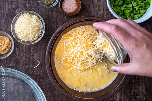 Ingredients for quinoa egg bake, raw egg mix in glass bowl with metal whisk, quinoa, cheese, herbs, broccoli, glass baking dish, on a wood table photo